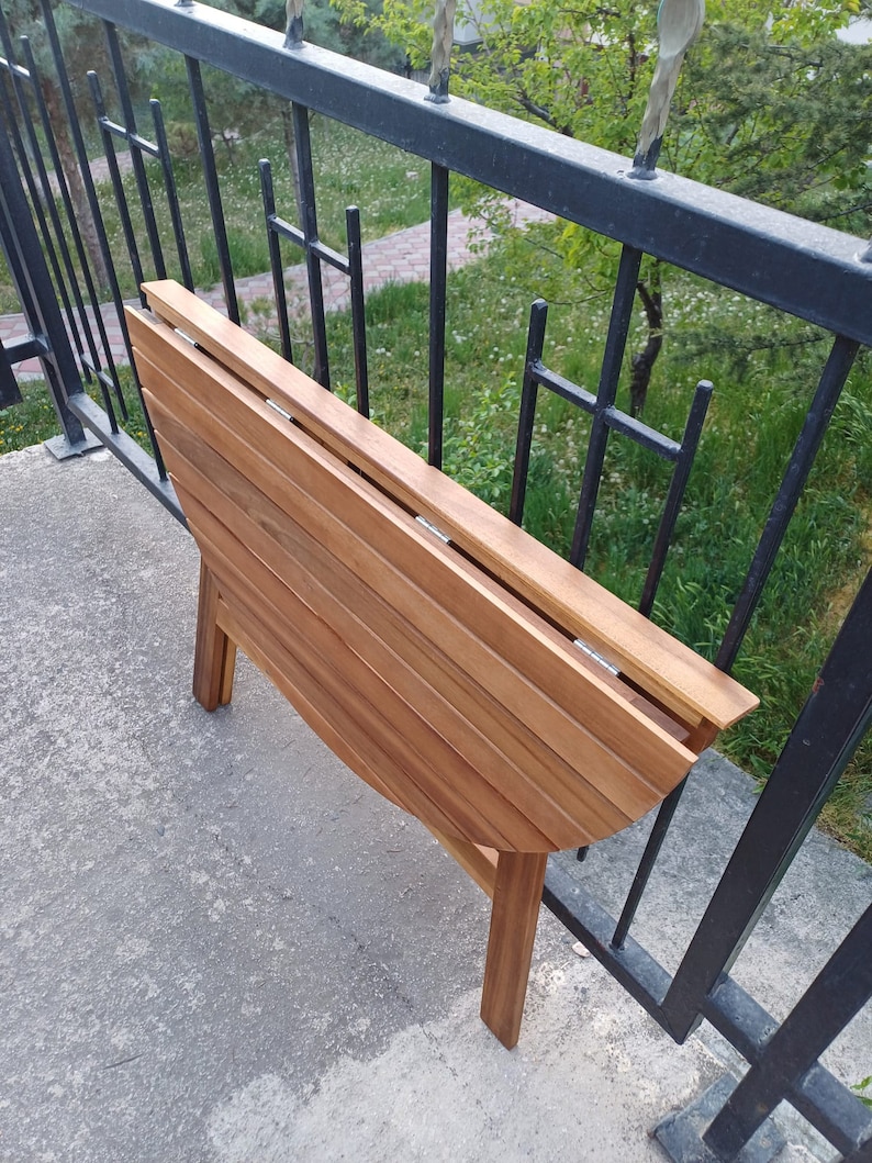 May include: A wooden folding table with a rounded top and a natural wood finish. The table is folded and standing on a concrete surface.