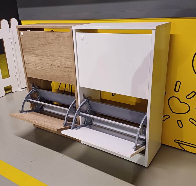 May include: Two shoe storage cabinets with flip-down doors. The cabinet on the left is wood-toned and the cabinet on the right is white. Both cabinets have a gray metal frame and a shelf.