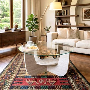 May include: A round glass coffee table with a light wood base featuring a curved design. The table holds a white teacup and saucer, a book with text on the cover, and a small gold-coloured metal motorcycle model on the lower shelf.