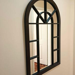 May include: An arched wall mirror with a black frame featuring a windowpane design. The mirror has a rounded top section with a semi-circular grid pattern and rectangular panes below, creating a classic architectural look for any interior space.