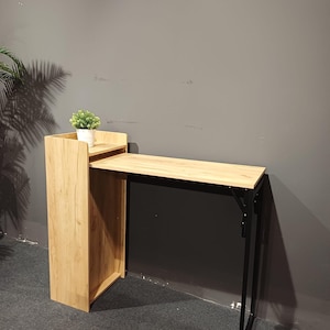 May include: A light wood-tone folding desk with a black metal frame. The desk has a small shelf on top with a small potted plant. The desk is designed to fold up for storage. The desk is 40 inches long.