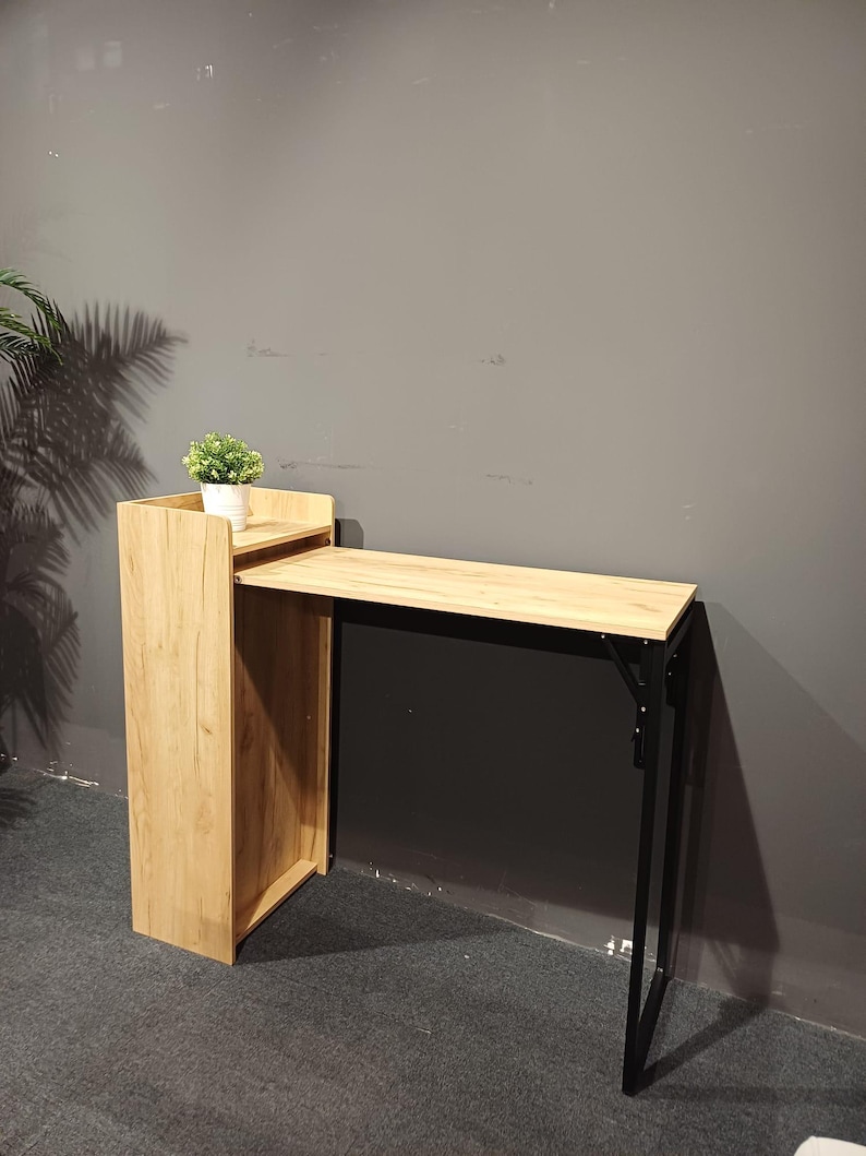 May include: A light wood-tone folding desk with a black metal frame. The desk has a small shelf on top with a small potted plant. The desk is designed to fold up for storage. The desk is 40 inches long.