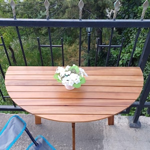 May include: A round wooden table with a natural wood finish and a white flower arrangement in a white pot. The table is on a balcony with a black metal railing and a view of green trees.
