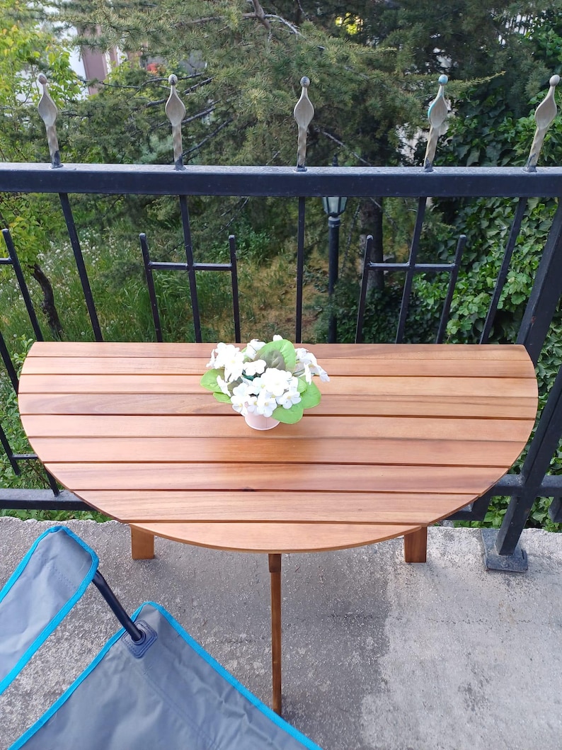 May include: A round wooden table with a natural wood finish and a white flower arrangement in a white pot. The table is on a balcony with a black metal railing and a view of green trees.