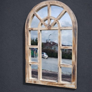 May include: An arched window-style wall mirror with a distressed, light-colored wooden frame featuring dark brown brush strokes. The frame is divided into multiple rectangular and curved panes, reflecting an outdoor scene with a car and buildings.