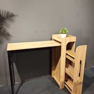 May include: A light wood-toned desk with a black metal frame and a matching side storage unit. A small potted plant sits on top of the storage unit. The desk is designed for compact spaces.