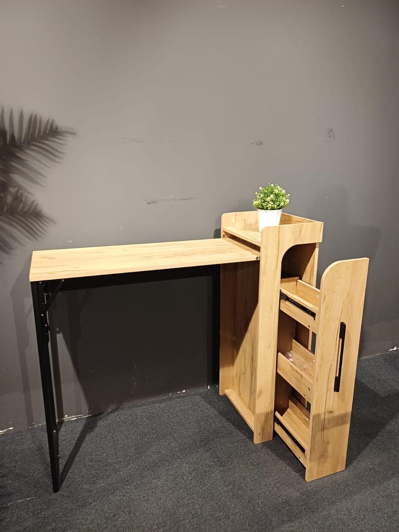 May include: A light wood-toned desk with a black metal frame and a matching side storage unit. A small potted plant sits on top of the storage unit. The desk is designed for compact spaces.