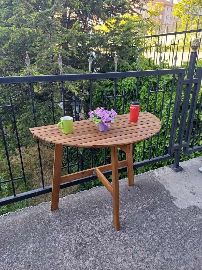 May include: A brown wooden half-round table with a green mug and a red travel mug on the surface. A potted plant with purple flowers sits on the table. The table is on a balcony with a black metal railing.