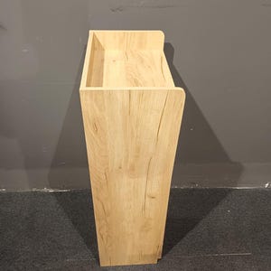 May include: A tall, light-colored wooden cabinet with a rectangular top and a tapered body. The cabinet has a natural wood grain finish and a simple, modern design. The top has a recessed area, and the cabinet is standing on a dark gray carpet.