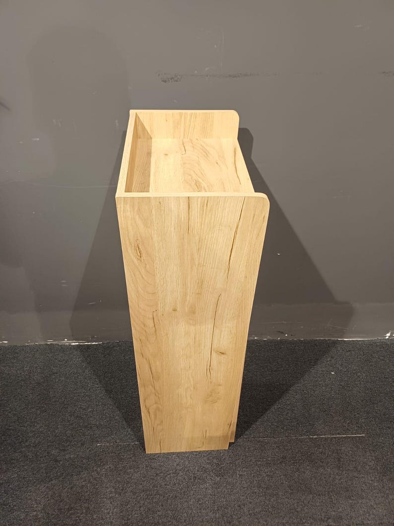 May include: A tall, light-colored wooden cabinet with a rectangular top and a tapered body. The cabinet has a natural wood grain finish and a simple, modern design. The top has a recessed area, and the cabinet is standing on a dark gray carpet.
