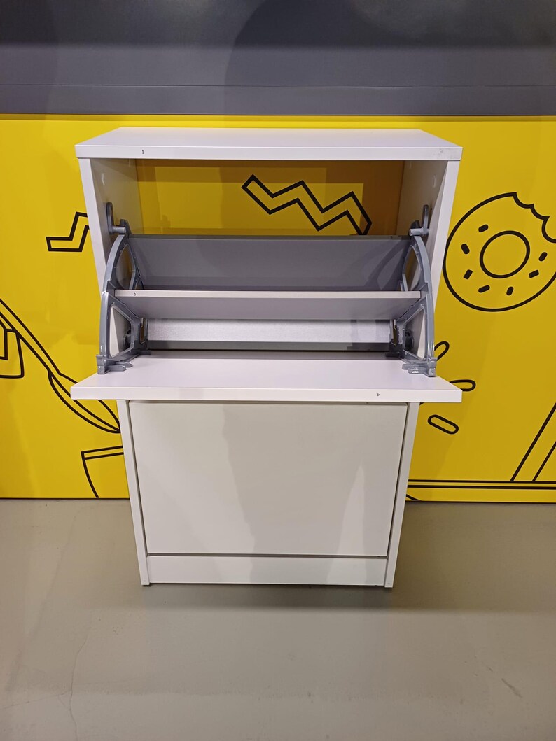 May include: White shoe storage cabinet with two shelves that fold down. The cabinet has a simple design and is made of wood.