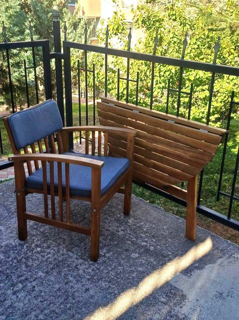 May include: A wooden folding table with a semi-circular top and a matching wooden chair with a blue cushion. The table and chair are on a gray concrete patio.