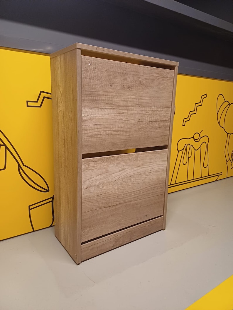 May include: A light brown wooden shoe cabinet with two shelves. The cabinet has a simple design and is made of wood.