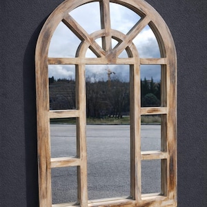 Puede incluir: Espejo de ventana arqueada de madera con acabado envejecido. El espejo refleja una escena exterior con una carretera, árboles y un cielo nublado. El marco tiene un diseño de rejilla con secciones rectangulares y arqueadas.