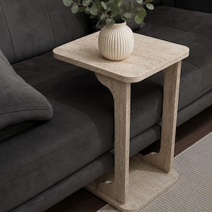 Puede incluir: Una mesa auxiliar cuadrada de color claro con un diseño único, con una encimera y una base que se desliza debajo de un sofá. Un pequeño jarrón blanco y acanalado con vegetación se encuentra sobre la mesa. La mesa es de material claro.