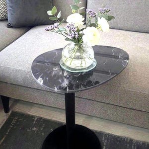 May include: A side table with an oval black marble top featuring white veining, supported by a single black pedestal base. A clear glass vase sits on the tabletop, containing a bouquet of white flowers and green eucalyptus leaves.