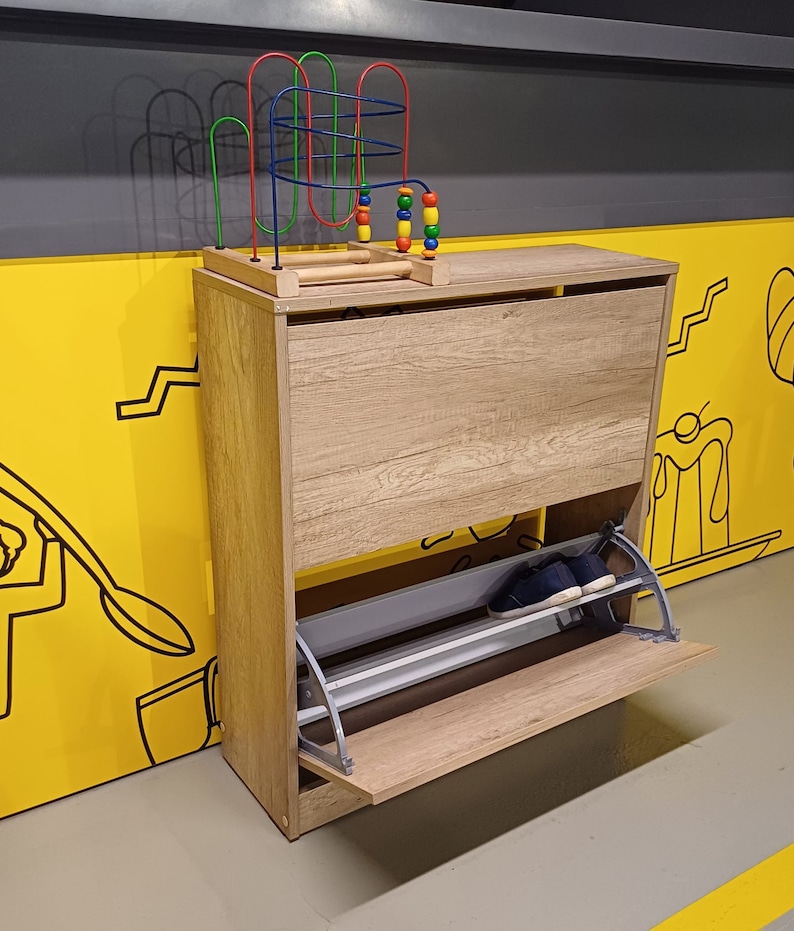 May include: A wooden shoe rack with a pull-out shelf. The shoe rack is light brown with a gray metal shelf. A colorful toy with beads is on top of the shoe rack.