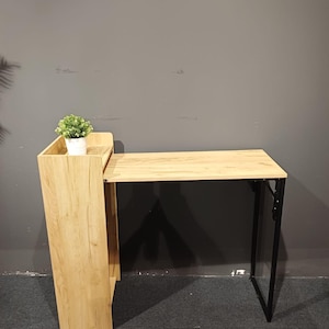 May include: A light wood-tone desk with a black metal frame. The desk has a built-in shelf and a small potted plant. The desk is designed to fold up for storage. The desk is suitable for a home office or small space.
