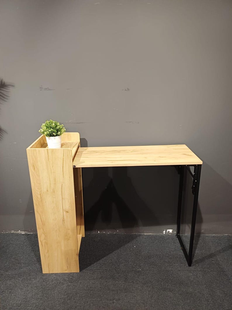 May include: A light wood-tone desk with a black metal frame. The desk has a built-in shelf and a small potted plant. The desk is designed to fold up for storage. The desk is suitable for a home office or small space.