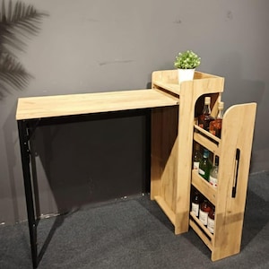 May include: A light wood-toned bar table with a black metal frame. The table has a built-in shelf with a small potted plant and a side rack for storing bottles. The table is designed for home use.
