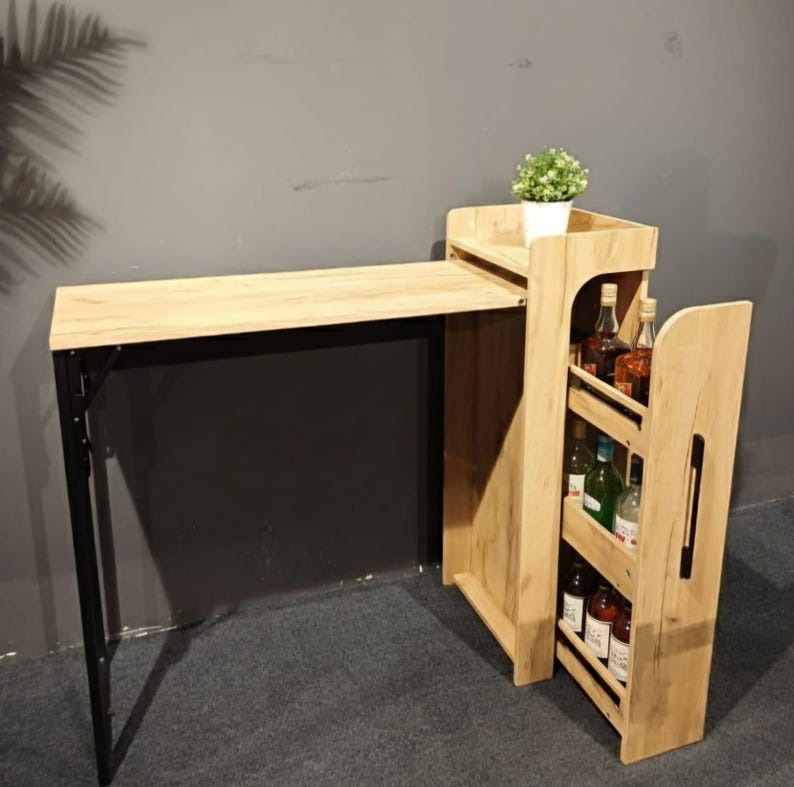 May include: A light wood-toned bar table with a black metal frame. The table has a built-in shelf with a small potted plant and a side rack for storing bottles. The table is designed for home use.