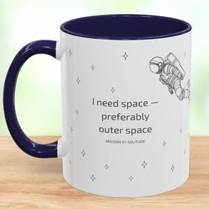 May include: A white ceramic mug with a dark blue interior and handle. The exterior features a black line drawing of an astronaut, small star icons, and the text: I need space — preferably outer space. MISSION 01: SOLITUDE is printed below.