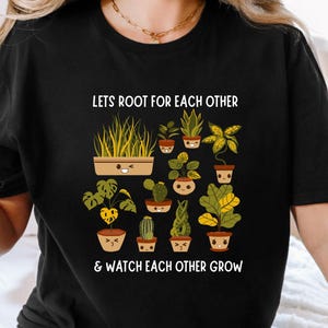 Könnte beinhalten: Schwarzes T-Shirt mit einer Grafik verschiedener Topfpflanzen mit lächelnden Gesichtern. Der Text "Lets Root For Each Other & Watch Each Other Grow" ist über und unter den Pflanzenillustrationen gedruckt.