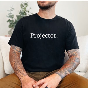 May include: A person wearing a black t-shirt with the word "Projector." printed in white on the front. The person is sitting in a chair and holding a glass of liquid.
