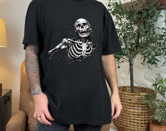 May include: A black short-sleeve t-shirt featuring a graphic print of a white skeleton torso emerging from a cracked surface. The skeleton is depicted with an open mouth and detailed rib cage, creating a high-contrast design on the dark fabric.