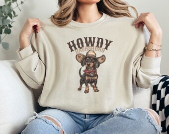 May include: A beige sweatshirt with the text "HOWDY DACHSHUND" in brown lettering. A cartoon dachshund wearing a cowboy hat and bandana is printed on the front of the sweatshirt.