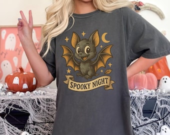 Puede incluir: Camiseta gris oscuro con un diseño de murciélago de dibujos animados, alas naranjas, estrellas y una luna creciente. El murciélago sonríe sobre una pancarta que dice "Spooky Night".