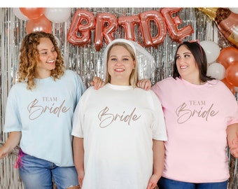 May include: Three women wearing matching t-shirts with the text "Team Bride" in rose gold lettering. The shirts are light blue, white, and pink. The women are smiling and posing for a photo.