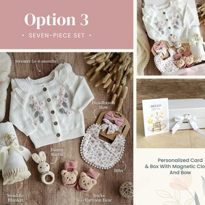 May include: A seven-piece baby gift set in a white box with a magnetic closure and a white bow. The set includes a white sweater with floral embroidery, a white swaddle blanket, a white bib with floral print, a pair of pink socks with a cartoon bear, a white headband with a pink bow, a bunny rattle, and a personalized card that says "Hello Little One".