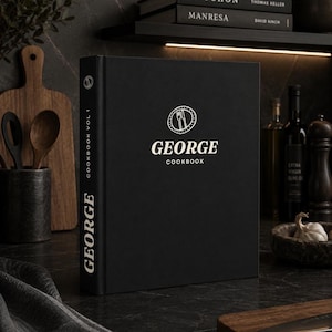 May include: A black hardback cookbook titled GEORGE COOKBOOK with white text and a circular logo featuring a fork and knife icon on the front cover. The spine of the book also displays the word GEORGE and the text COOKBOOK VOL 1 in white.