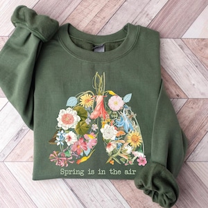 Puede incluir: Una sudadera verde oliva con un diseño floral de pulmones, llena de varias flores coloridas. El texto "Spring is in the air" está impreso en la parte inferior. La sudadera está hecha de un material suave y tiene mangas largas.