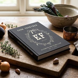 Personalized recipe book for couples in pepper black linen hardcover, monogram EF inside a hand drawn botanical wreath, Cook Together above and the names Elizabeth and Fitzwilliam with their wedding date below, spine and back cover shown.