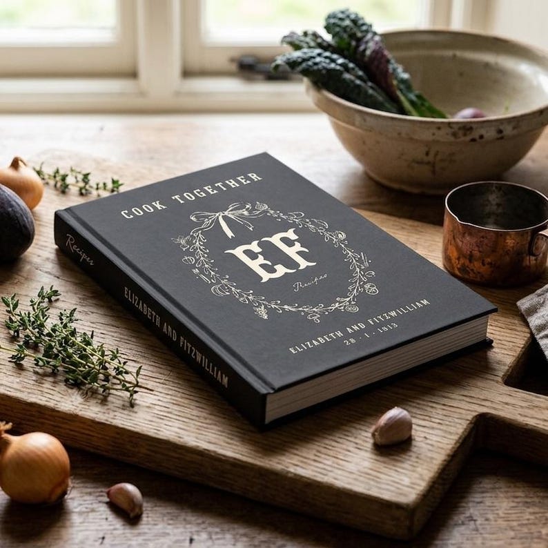 Personalized recipe book for couples in pepper black linen hardcover, monogram EF inside a hand drawn botanical wreath, Cook Together above and the names Elizabeth and Fitzwilliam with their wedding date below, spine and back cover shown.