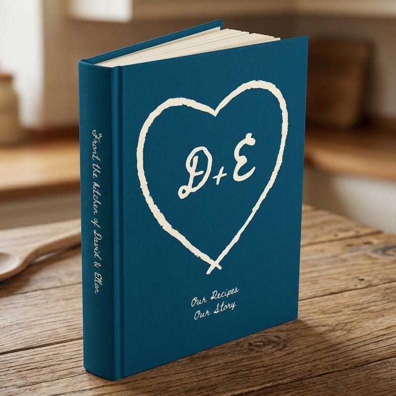 Personalized recipe book for couples in marine hardcover, a hand drawn carved heart with the initials D and E, Our Recipes Our Story below, From the kitchen of David and Elton on the spine and made with love on the back.