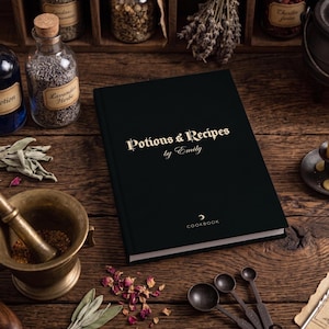Puede incluir: Un libro de tapa dura negra con el texto dorado Potions & Recipes by Emily y la palabra COOKBOOK impresa en la portada. El libro está colocado sobre una superficie de madera junto a un mortero de latón, cucharas medidoras y frascos de vidrio.