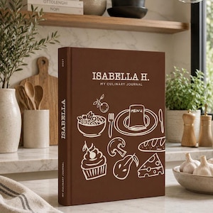 May include: A brown hardback culinary journal with the text ISABELLA H. MY CULINARY JOURNAL on the front cover. The cover features white line art illustrations of a bowl, a plate with a menu, a cupcake, a mushroom, a pear, and a piece of cheese.