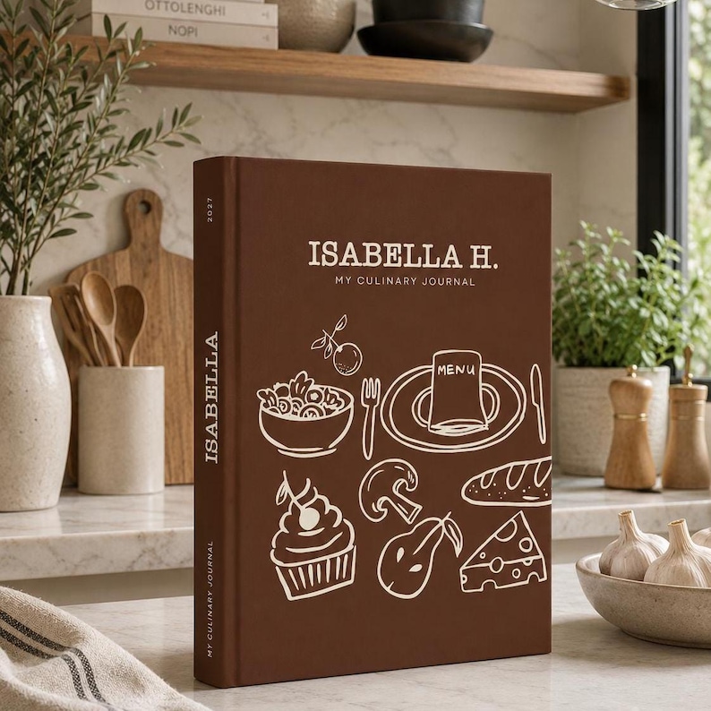 May include: A brown hardback culinary journal with the text ISABELLA H. MY CULINARY JOURNAL on the front cover. The cover features white line art illustrations of a bowl, a plate with a menu, a cupcake, a mushroom, a pear, and a piece of cheese.