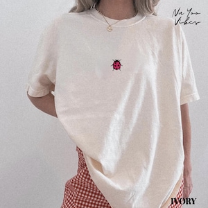 Könnte beinhalten: Elfenbeinfarbenes T-Shirt mit einem kleinen, gestickten Marienkäfer-Design. Das Shirt hat kurze Ärmel und eine lockere Passform. Die Worte "Nu Yoo Vibes" sind in handschriftlicher Form auf der oberen rechten Seite des Shirts aufgedruckt. Das Wort "IVORY" ist unten rechts aufgedruckt.