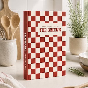 May include: A hardback recipe book featuring a red and white chequered pattern on the cover and spine. The text on the front and spine reads, FROM THE KITCHEN OF THE GREEN'S, printed in a bold, dark red font against a white rectangular background.
