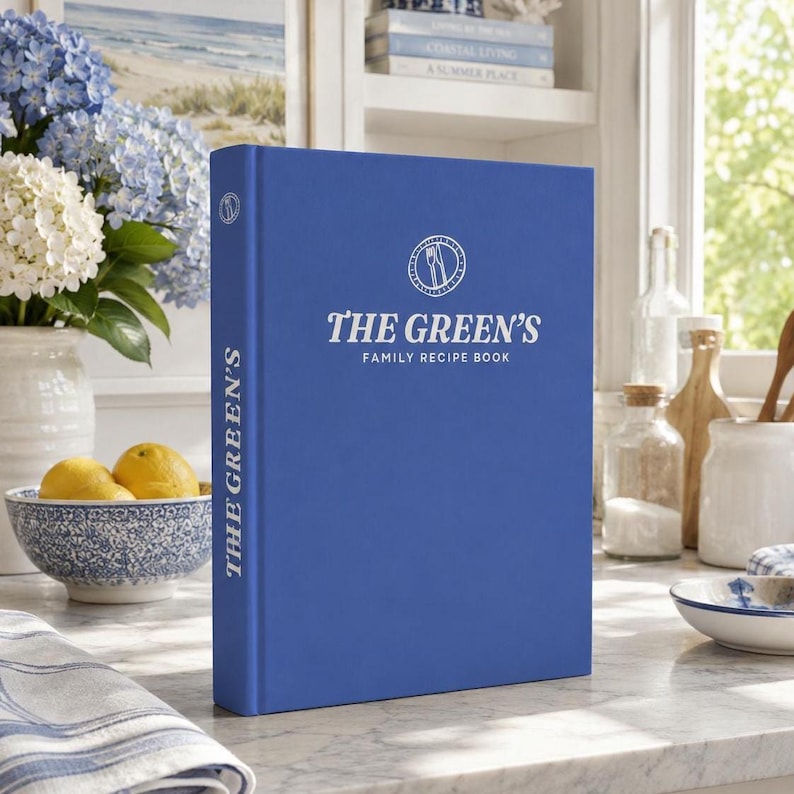 May include: A vibrant blue hardback book titled The Green's Family Recipe Book, featuring a white circular emblem with a fork and knife icon on the front cover and the title printed vertically in white text on the spine of the book.