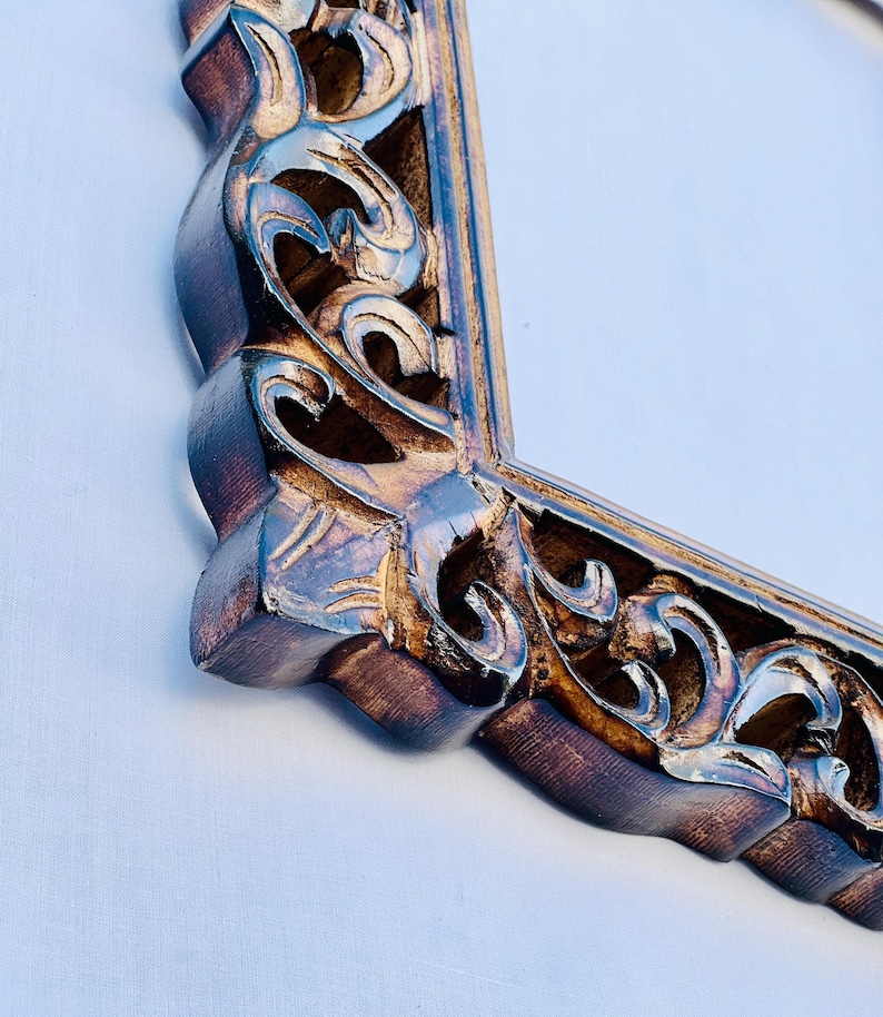 May include: A close-up of a wooden mirror frame with intricate carvings. The frame is a dark brown color with gold accents. The mirror is rectangular and has a beveled edge.