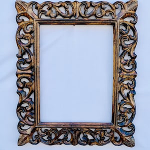 May include: A large, ornate wooden picture frame with intricate carvings. The frame is a dark brown color with a slightly reddish hue. The frame is rectangular and has a slightly distressed look.