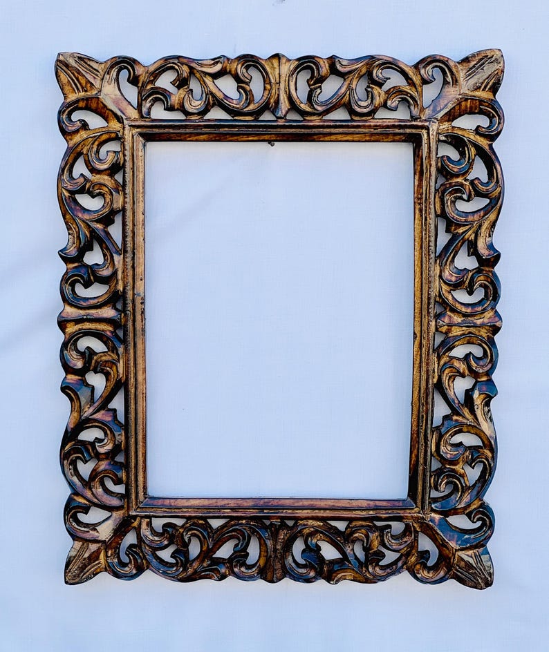 May include: A large, ornate wooden picture frame with intricate carvings. The frame is a dark brown color with a slightly reddish hue. The frame is rectangular and has a slightly distressed look.
