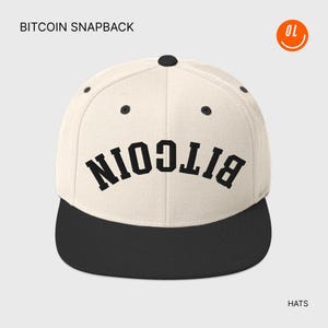 May include: A cream-colored snapback hat with a black brim. The word "BITCOIN" is printed in large, black, block letters across the front. The hat has a flat brim and a button on top. The word "HATS" is printed at the bottom.