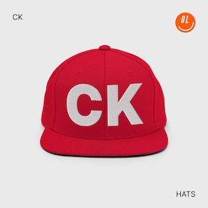 Peut inclure: Casquette rouge à visière plate avec les lettres blanches "CK" brodées sur le devant. Le chapeau a une couronne structurée et une visière plate. Le mot "HATS" est en bas à droite.