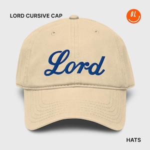 May include: A beige baseball cap with the word "Lord" embroidered in blue cursive script across the front. The hat has a curved brim and a six-panel design. The text "LORD CURSIVE CAP" is above the hat, and "HATS" is below.